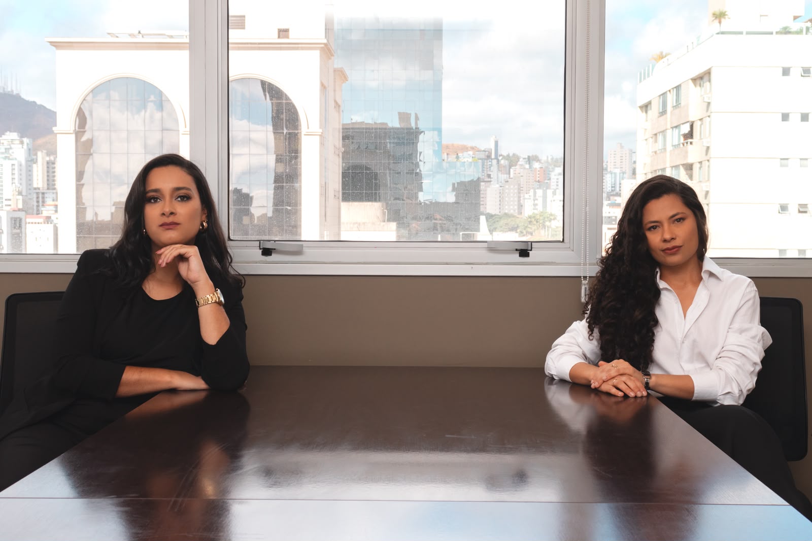 As advogadas Camila Miranda e Ana Flávia Barbosa sentadas à mesa de reunião, com a vista de Belo Horizonte ao fundo